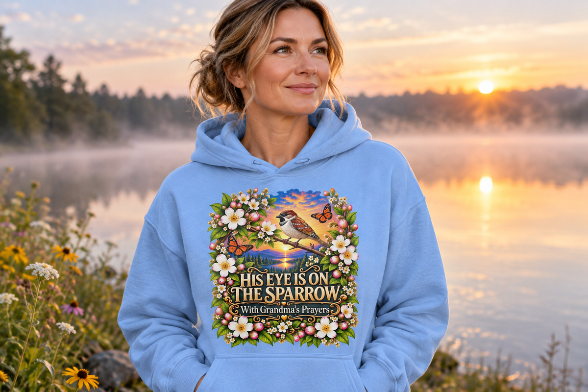 Adult woman wearing the powder-blue His Eye Is on the Sparrow hoodie beside a misty lakeshore at sunrise