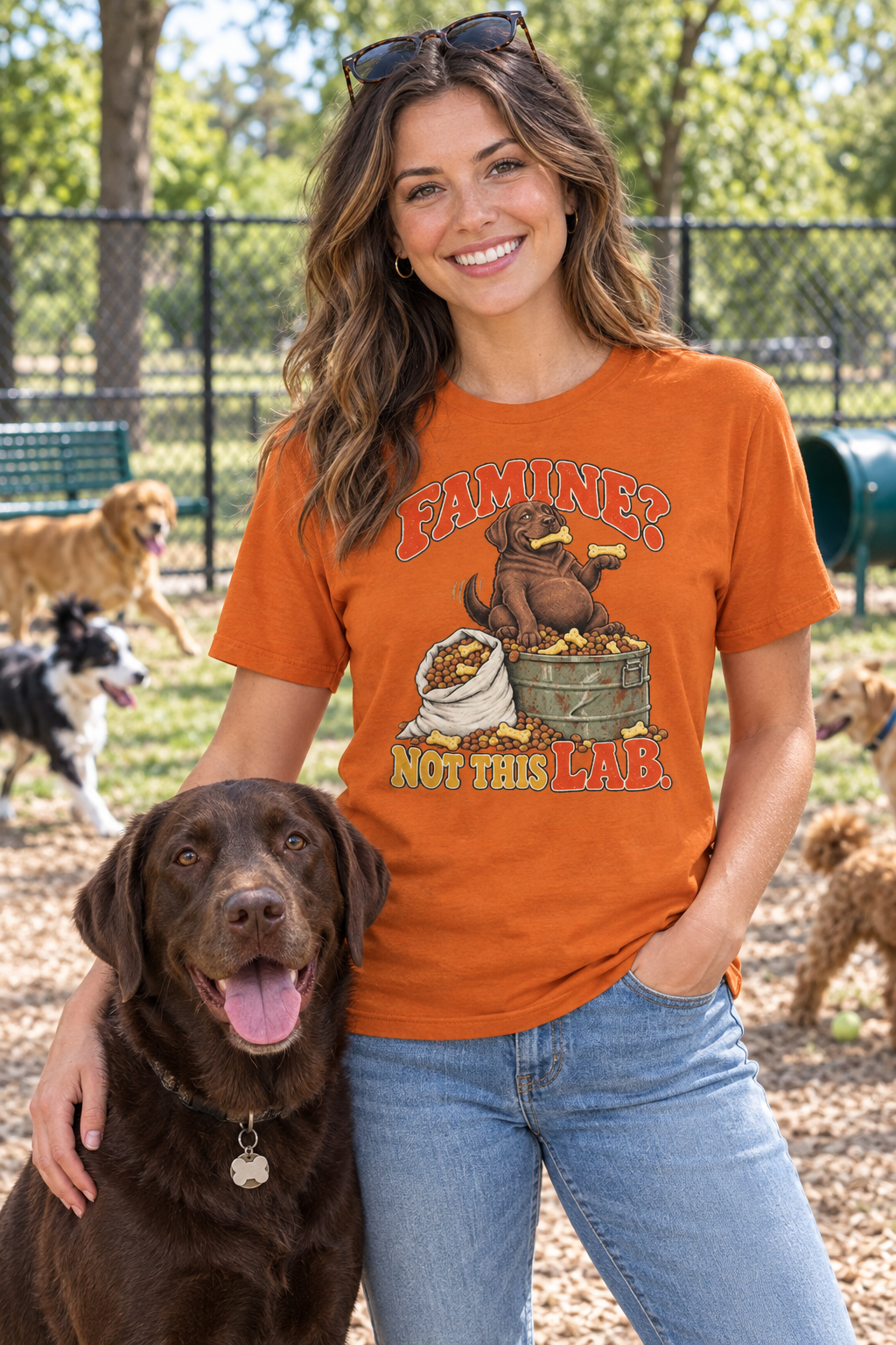 Woman wearing an orange funny Labrador graphic T-shirt with dogs at a dog park